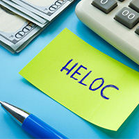 HELOC written on yellow sticky note with $100 bills, calculator, and pen on blue background.