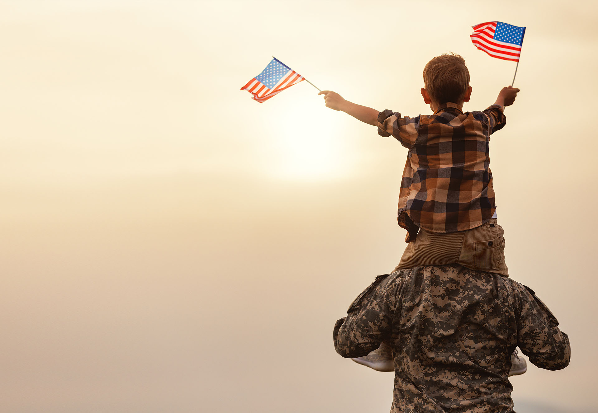 Image of back of military father holding son on shoulders waving American flags.