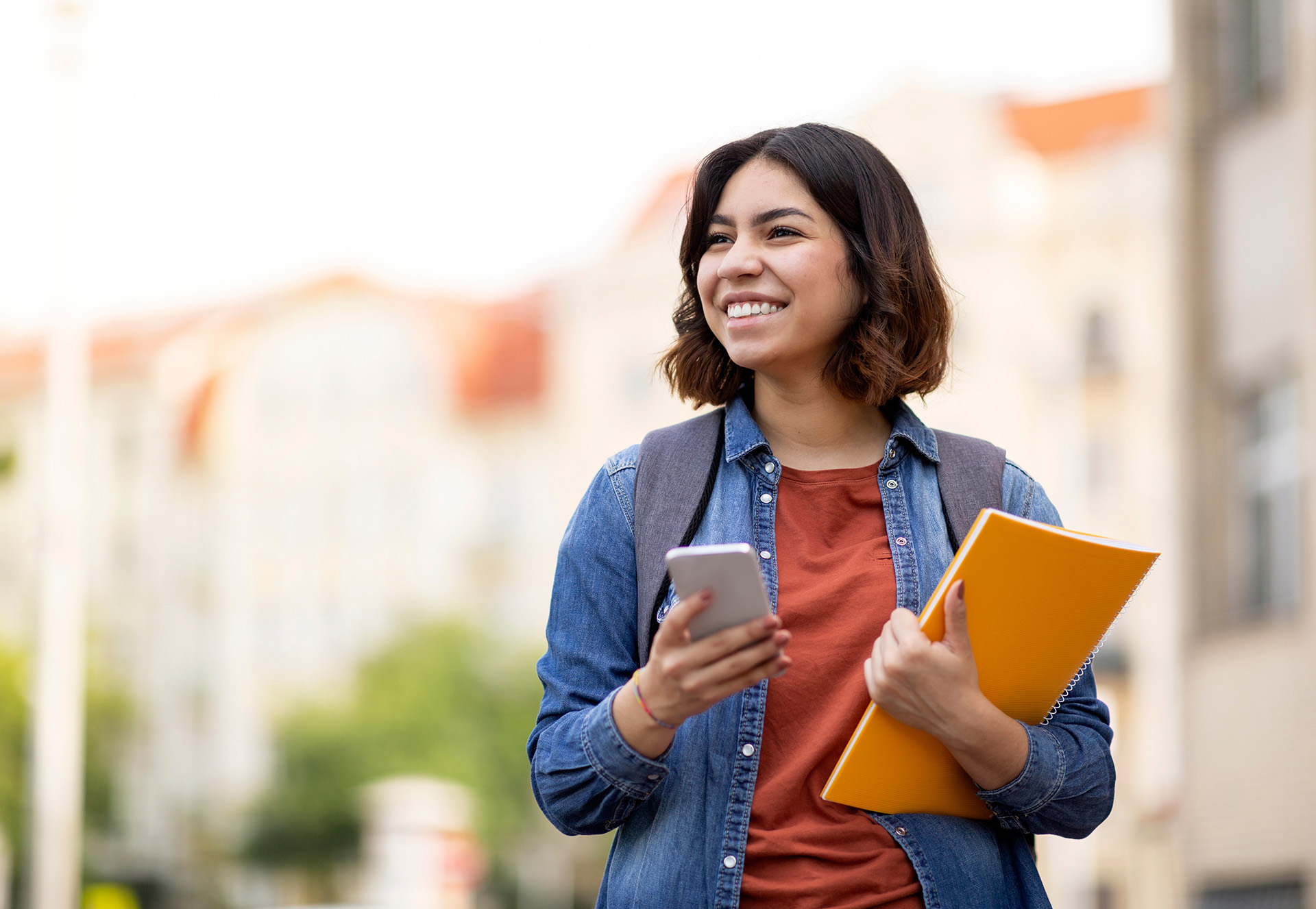 Cheerful middle eastern female student with smartphone and workbooks standing outdoors.